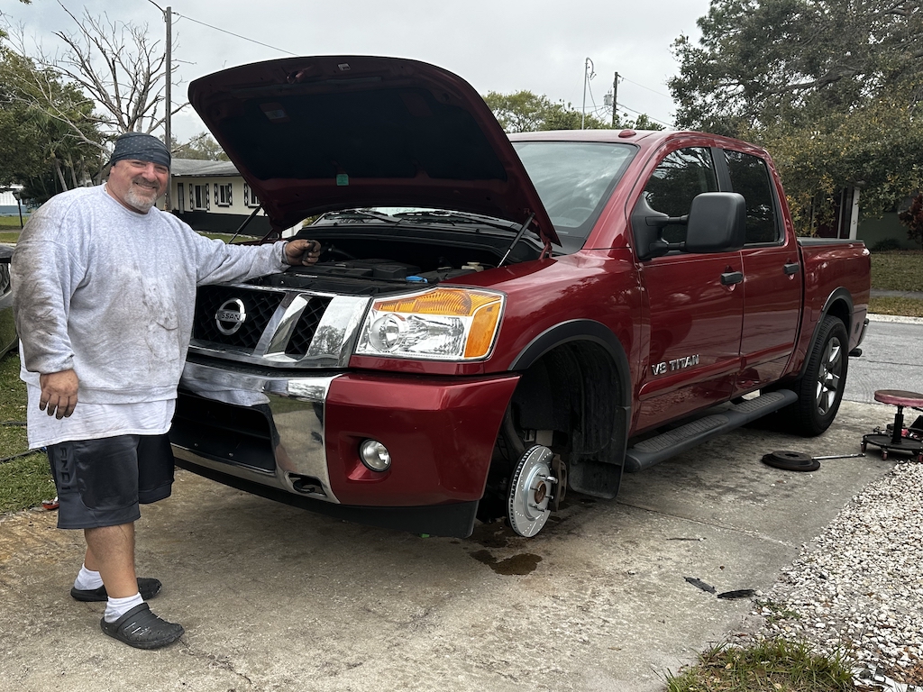 Bob Pacecca, professional mobile mechanic, standing next to a red Nissan Titan pickup truck with hood open, demonstrating mobile automotive repair services in a residential driveway setting